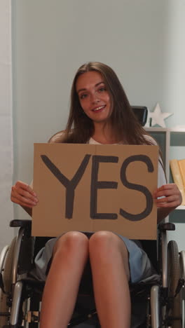 happy woman sits in wheelchair near houseplant holding poster with word yes. young person with disability with paralysis smiles looking in camera