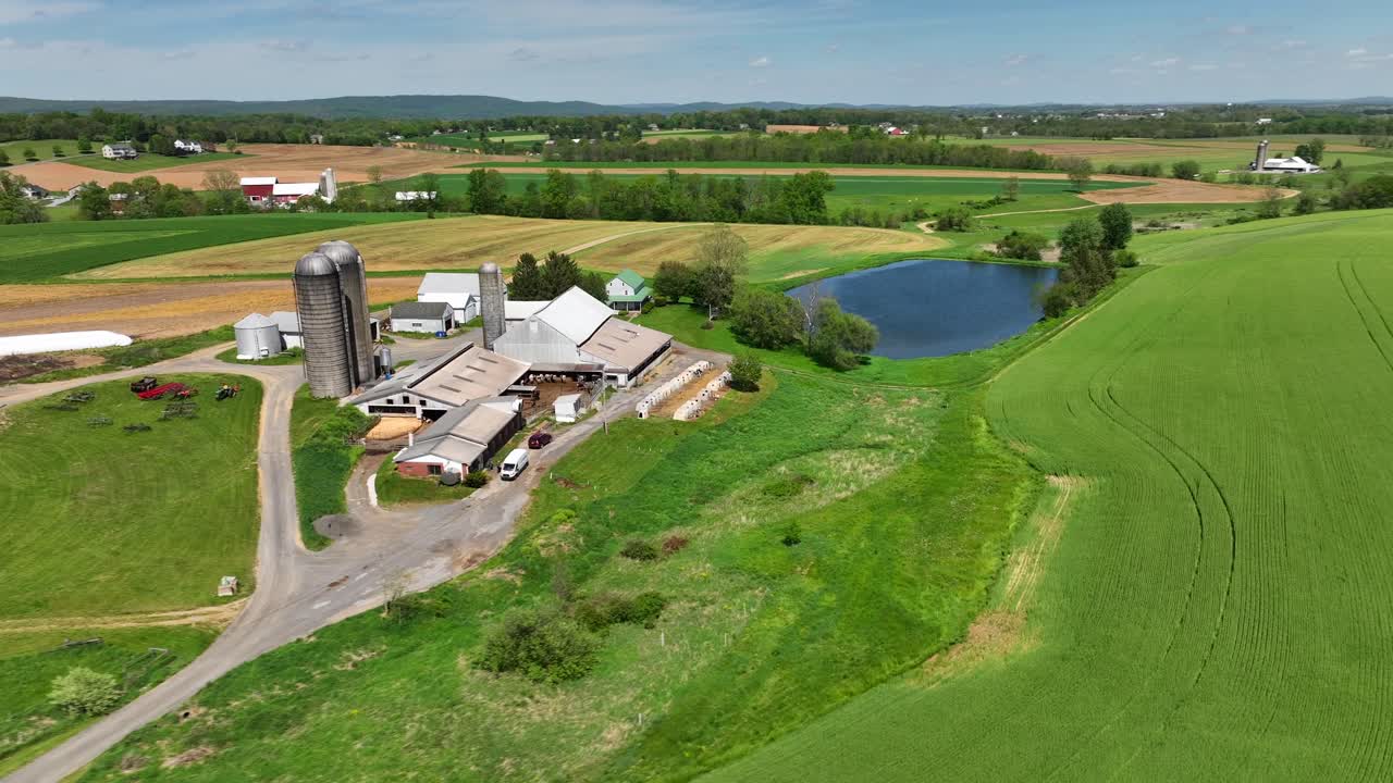 vista aérea de una serena granja rural con silos, graneros, un estanque y vastos campos verdes, rodeados de colinas onduladas y granjas lejanas