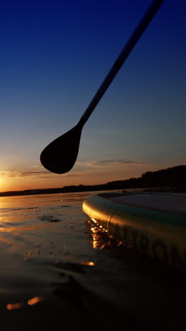 male silhouette approaching a camera on sup board. man uses an oar to push and move past camera. low angle view. vertical video.