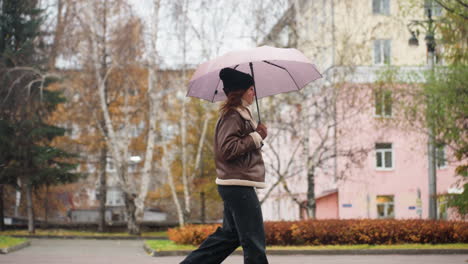 lady running happily in light snowfall holding umbrella wearing knit cap brown shearling jacket black trousers one hand pocket surrounded by colorful autumn leaves in calm urban setting