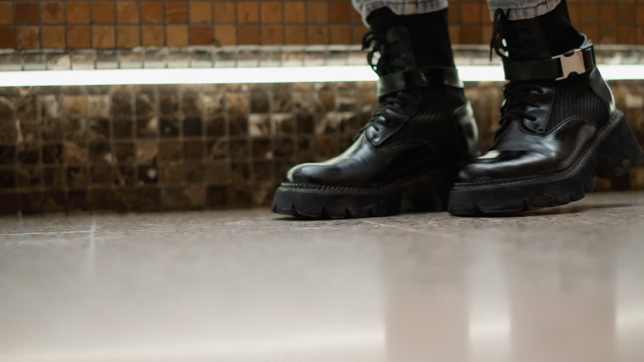 Low angle close up of stylish woman in black leather boots stepping onto mosaic bench seat near flowing mall fountain, showing leg motion and textured tile background in modern indoor setting
