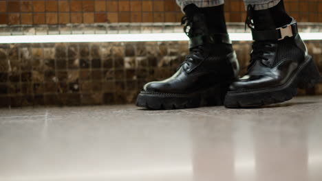 low angle close up of stylish woman in black leather boots stepping onto mosaic bench seat near flowing mall fountain, showing leg motion and textured tile background in modern indoor setting