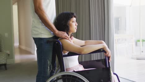 Biracial-woman-in-wheelchair-and-male-partner-holding-hands-by-window-in-sunny-living-room