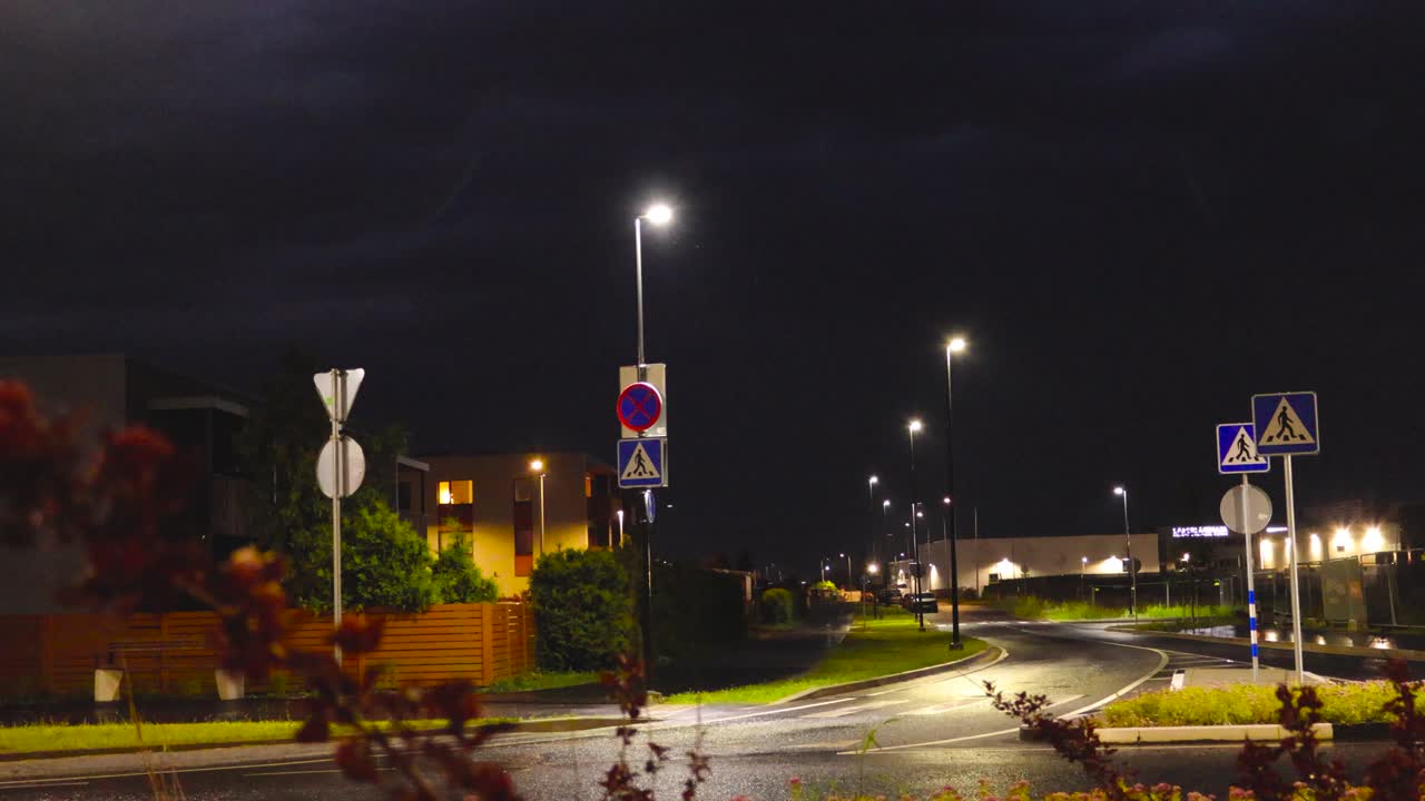 Static nighttime shot on a quiet suburban street while flashes of lightning in the night sky. Forks of lightning flash across the dark dense storm clouds. Streetlights illuminate the wet asphalt road