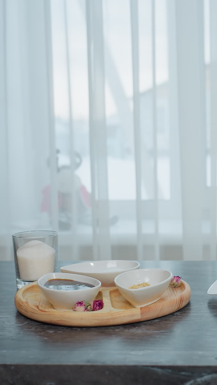 elegante mesa de cocina negra con bandeja de madera que contiene platos, especias y herramientas de cocina, libro de portada marrón colocado al lado de la mantequilla en el plato, cortina blanca transparente revela el edificio exterior a través de la ventana