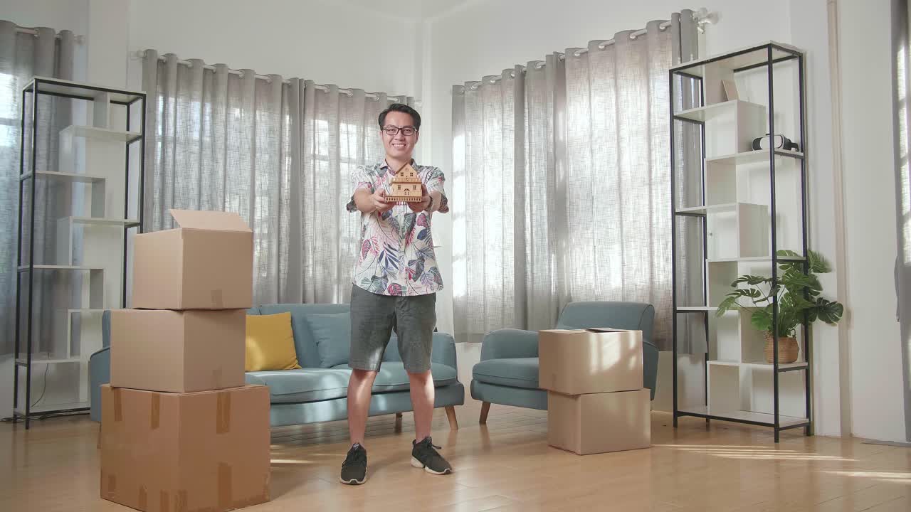 카드보드 상자를 가진 아시아인 남자가 미소 짓고 새로운 집에서 카메라에 집 모델을 보여줍니다.