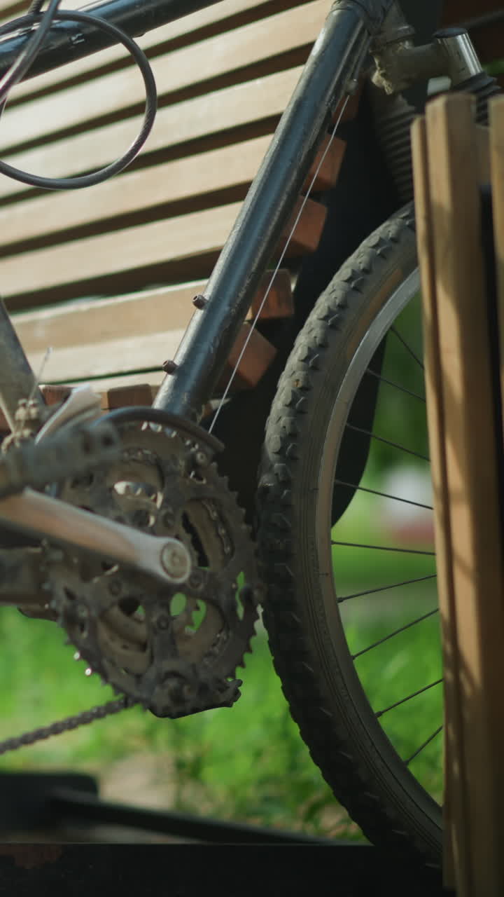 primer plano de una bicicleta colocada entre un banco de madera y una estructura cercana, descansando contra el banco mientras el ciclista se sienta, se inclina hacia atrás, descansando las manos en las piernas, rodeado de árboles verdes exuberantes al aire libre