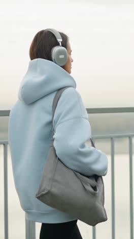 backside view of female adult walking beside iron rail, holding a bag, with river in the background, the scene evokes tranquility and peacefulness as she strolls along the railing near water