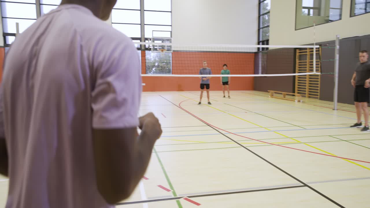 Diverse males watching man in pink shirt picking up volleyball and serving, starting rally in gym