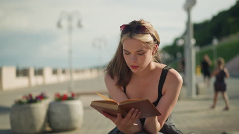 dama de soltera negra se sienta al aire libre, leyendo un libro cubierto de marrón mientras el viento voltea las páginas ligeramente, expresión pensativa en su rostro con un fondo borroso de personas que pasan