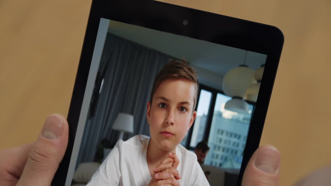 Primo piano di un bambino che parla tramite tablet durante una videochiamata con uno sconosciuto a casa. Padre che tiene in mano il tablet