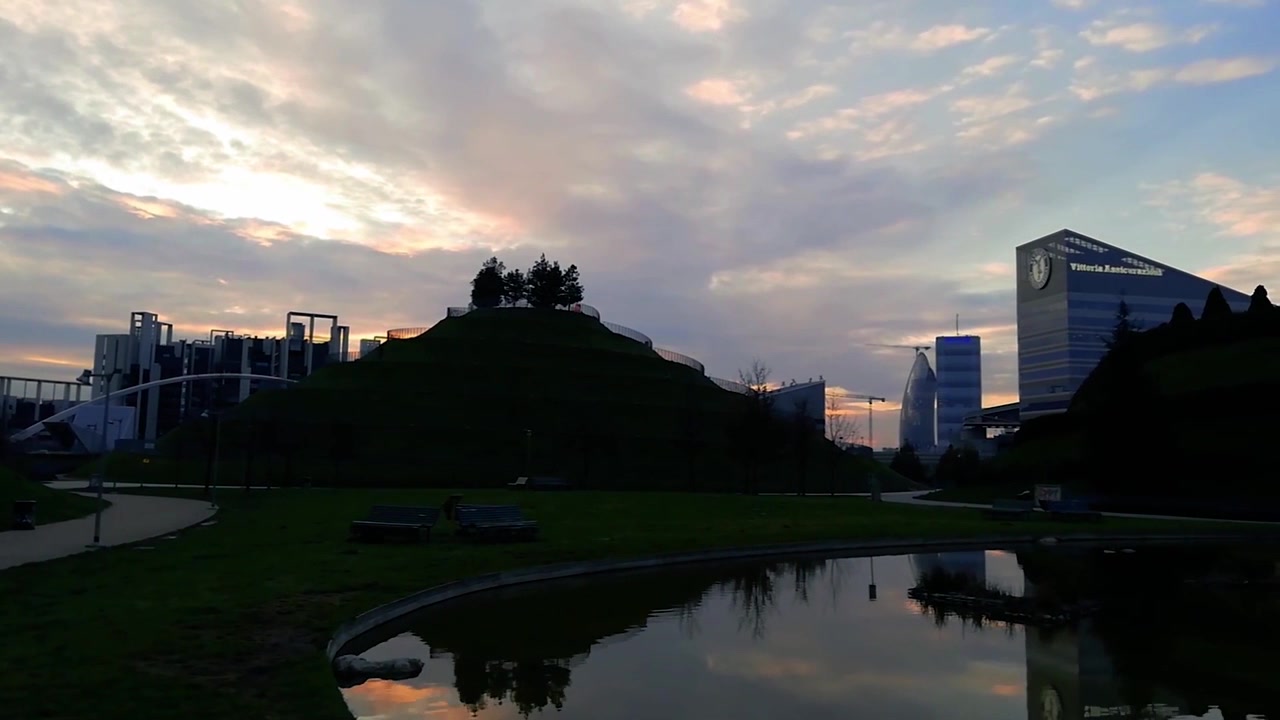 Parco Biblioteca degli Alberi em Milão, Itália, mostrando lago, colina gramada, trilhas para caminhada, edifícios modernos e céu noturno perto da Porta Nuova.