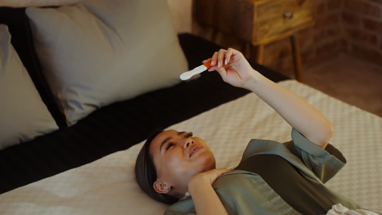 침대에 누워있는 여성이 임신 테스트를 보고 있습니다.