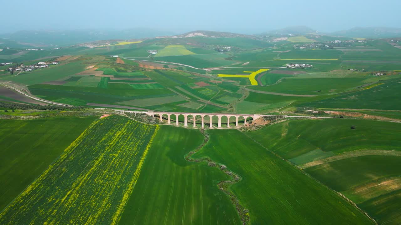 The scene is an aerial view of a bridge crossing over a valley with a train track running through it. On the other side of the valley, there are green fields and mountains in the distance.
