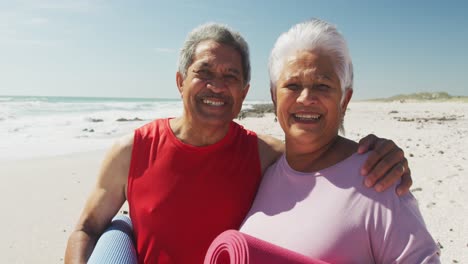 Portrait-of-hispanic-senior-couple-standing-on-beach,-holding-yoga-mat-and-smiling