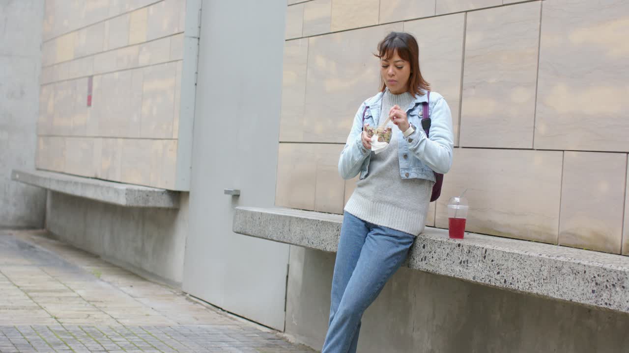Mujer asiática apoyada en un banco en la plaza, comiendo el almuerzo de un recipiente para llevar y bebiendo una bebida roja.