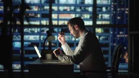 tired young man working on a laptop late night in the office. sleepy businessman sitting at desk in dark office. tired and stressed businessman in glasses works on a laptop of the night city office