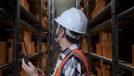 close up side view of asian male engineer with safety helmet using smartphone and looking around while standing in the warehouse with shelves full of delivery goods