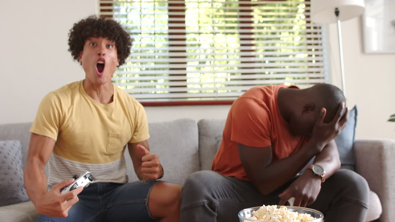 Playing video games, two multiracial male friends sitting on couch with bowl of popcorn, at home