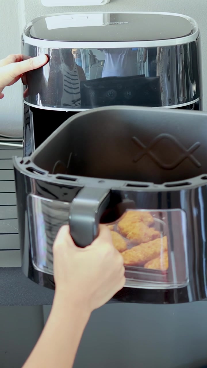 Una persona rimuove un cestino di cibo impanato cotto da una friggitrice ad aria da banco e lo posiziona su un tavolo da pranzo illuminato dal sole. Illuminazione luminosa e naturale, ripresa a mano.