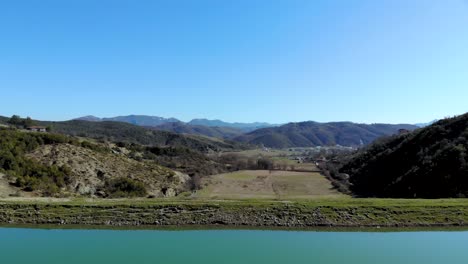 Calm-glassy-water-surface-of-reservoir-with-dam-over-farm-fields-and-village-houses-on-beautiful-valley-surrounded-by-mountains,-clear-blue-sky
