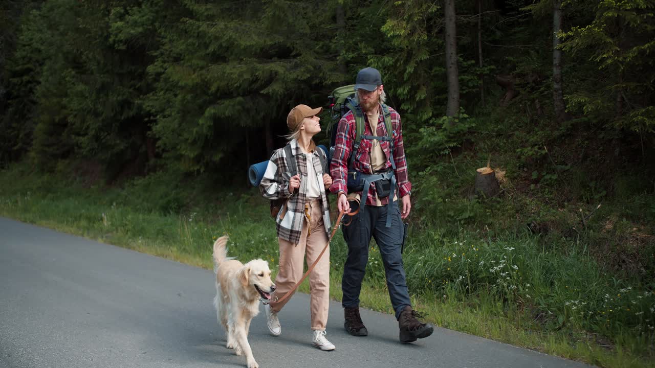 un chico y una chica viajeros con ropa especial para hacer senderismo caminan con su perro y colores claros a lo largo de la carretera a lo largo del bosque