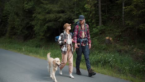 a guy and a girl travelers in special clothes for hiking walk with their dog and light coloring along the road along the forest