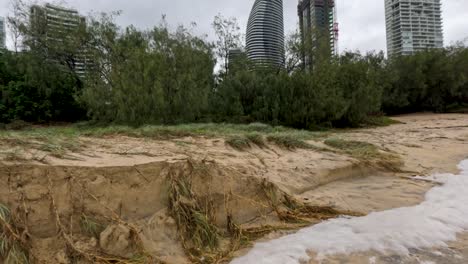 a beach shows severe erosion and pollution effects following cyclone alfred, with urban buildings in the background under cloudy skies