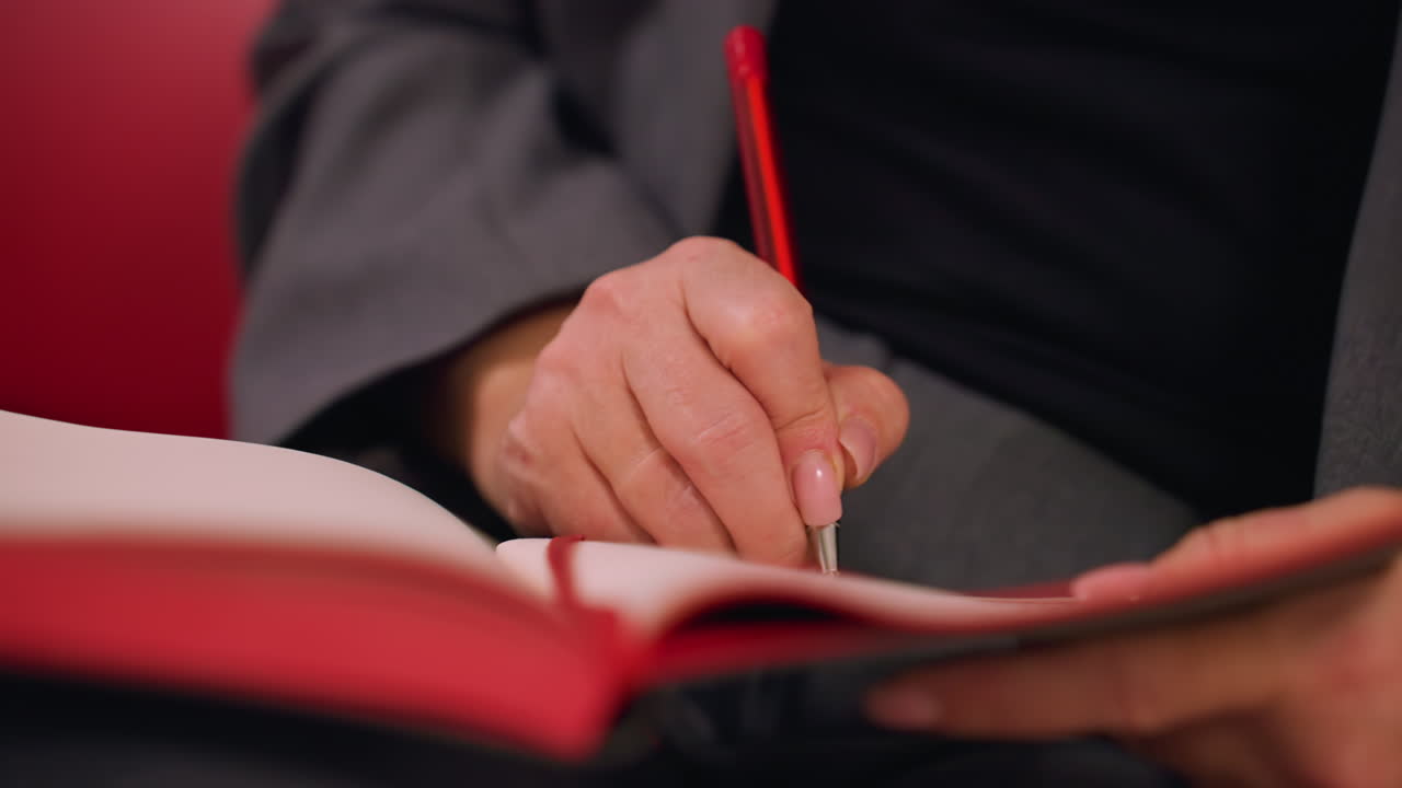 Close-up of person writing in red notebook with red pen, detailed hand and writing motion, focused on business or planning. Professional office setting, workspace, creativity, and productivity in action