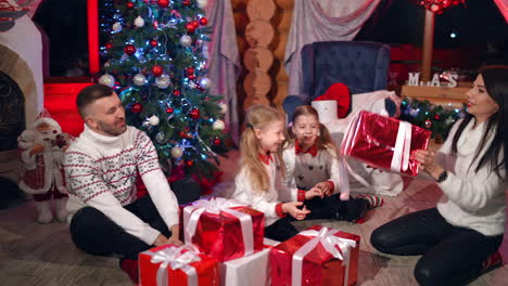 happy kids with presents at christmas. young family sitting together near beautiful christmas tree and holding bright red gift boxes with white ribbons. new year.