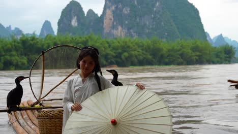 Hanfu-girl-holding-an-umbrella-poses-on-bamboo-raft-on-river-with-two-cormorants