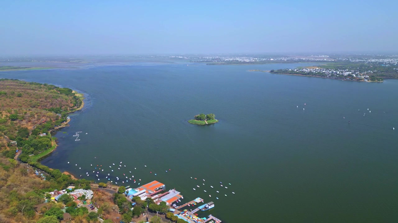 Riprese aeree con drone che sorvolano il Lago Superiore a Bhopal, con una piccola isola, acque estese, strutture sul lago e la costa della città in lontananza sotto cieli diurni sereni.
