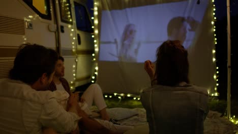 group of young cheerful people watching a movie on camping site on projection screen.