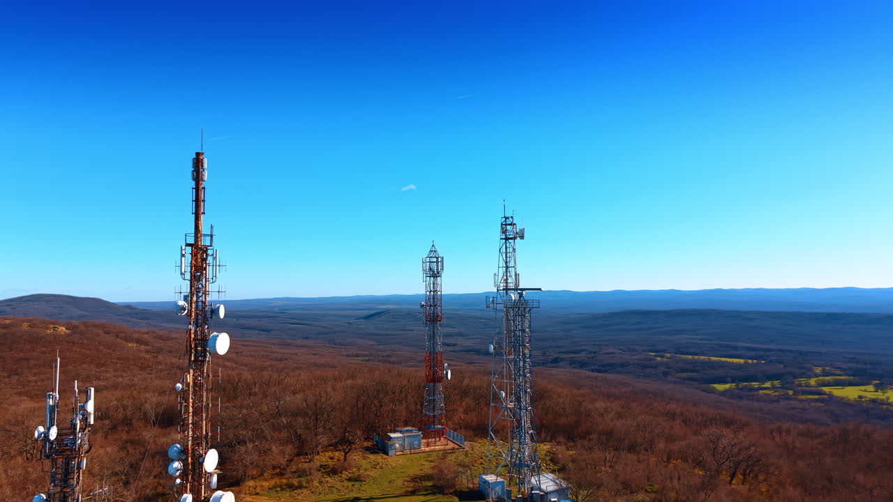 Telecommunication towers on forested hill under clear blue sky. Aerial shot of several cell towers and antennas on a ridge surrounded by autumn forest