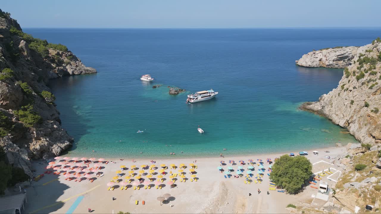 Parallel drone footage of Achata Beach in Karpathos, with umbrellas, sunbeds, swimmers and two tourist boats anchored on the rocks offshore in the turquoise Aegean waters