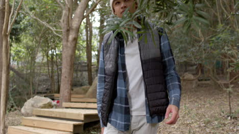 young man in forest wearing beanie and vest, looking up at trees