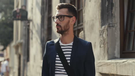 portrait of an attractive young man in glasses and stylish outfit turning his head to the camera and smiling in an old town street