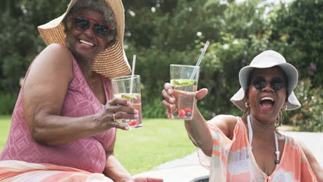 Retrato-De-Amigas-Afroamericanas-Mayores-Con-Sombreros-Para-El-Sol-Haciendo-Un-Brindis-En-El-Jardín,-Cámara-Lenta