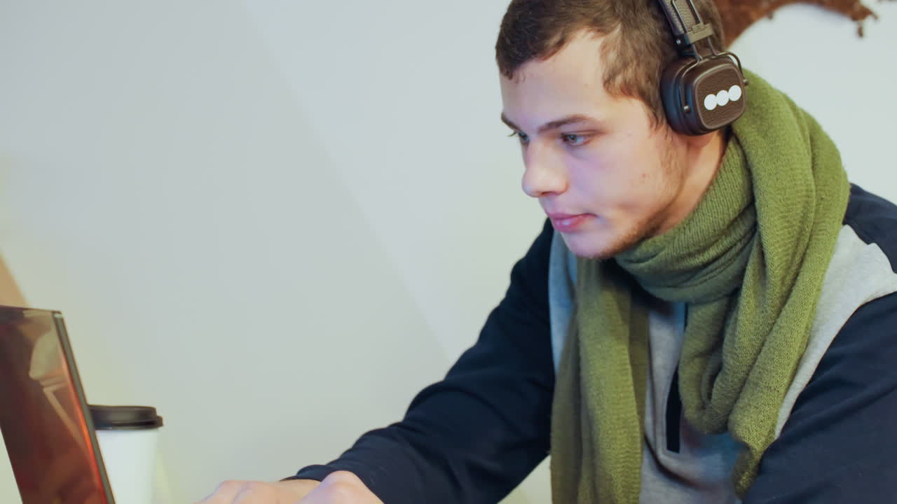 Jovem branco com fones de ouvido estudando em canto de estudo iluminado, cachecol verde, laptop e xícara de café, close-ups de perfil perto da estante e da xícara, expressão focada com as mãos no teclado.