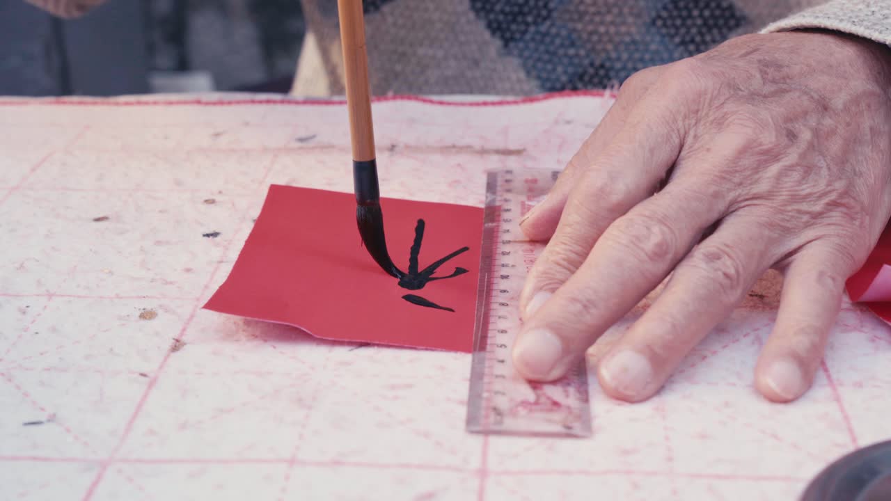 Close up static shot of an elderly person writing Chinese calligraphy with a brush on red paper using a brush aruler as guide on a traditional practice mat indoors with focused and cultural atmosphere