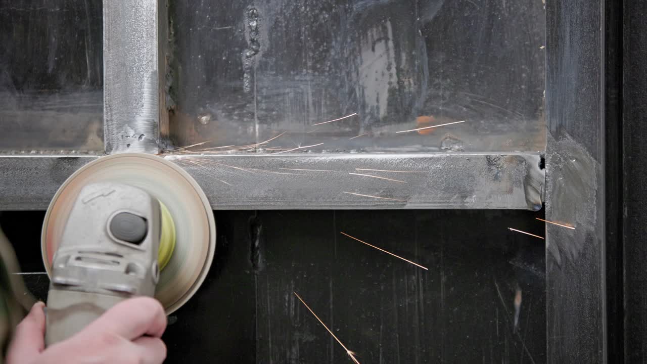 코카시안 손으로 용접 된 접합을 청소하는 글 그린더와 플래프 디스크로 용접 된 금속 구조에 긴 스파크 트레일과 함께 클로즈업, 느린 동작