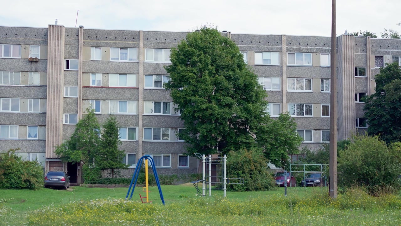 Bredt statisk skud af sovjettidens betonboligblok med legeplads i grøn gårdhave i Daugavpils, Letland, på en fredelig sommerdag