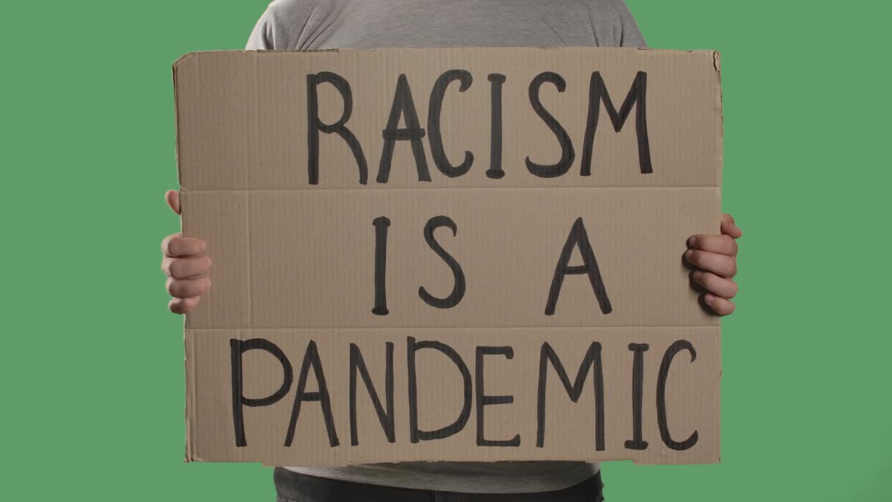 A man holds in front of him a poster from a cardboard box with the words RACISM IS A PANDEMIC. Isolated a green screen, chroma key. Close up. Slow motion