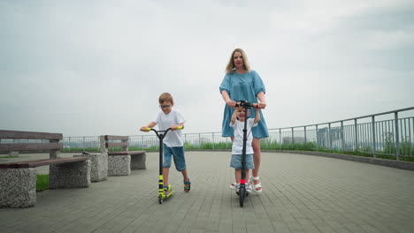 a mother and her children are riding scooters along a curved pathway, the younger child is with his mum, while the older child rides independently