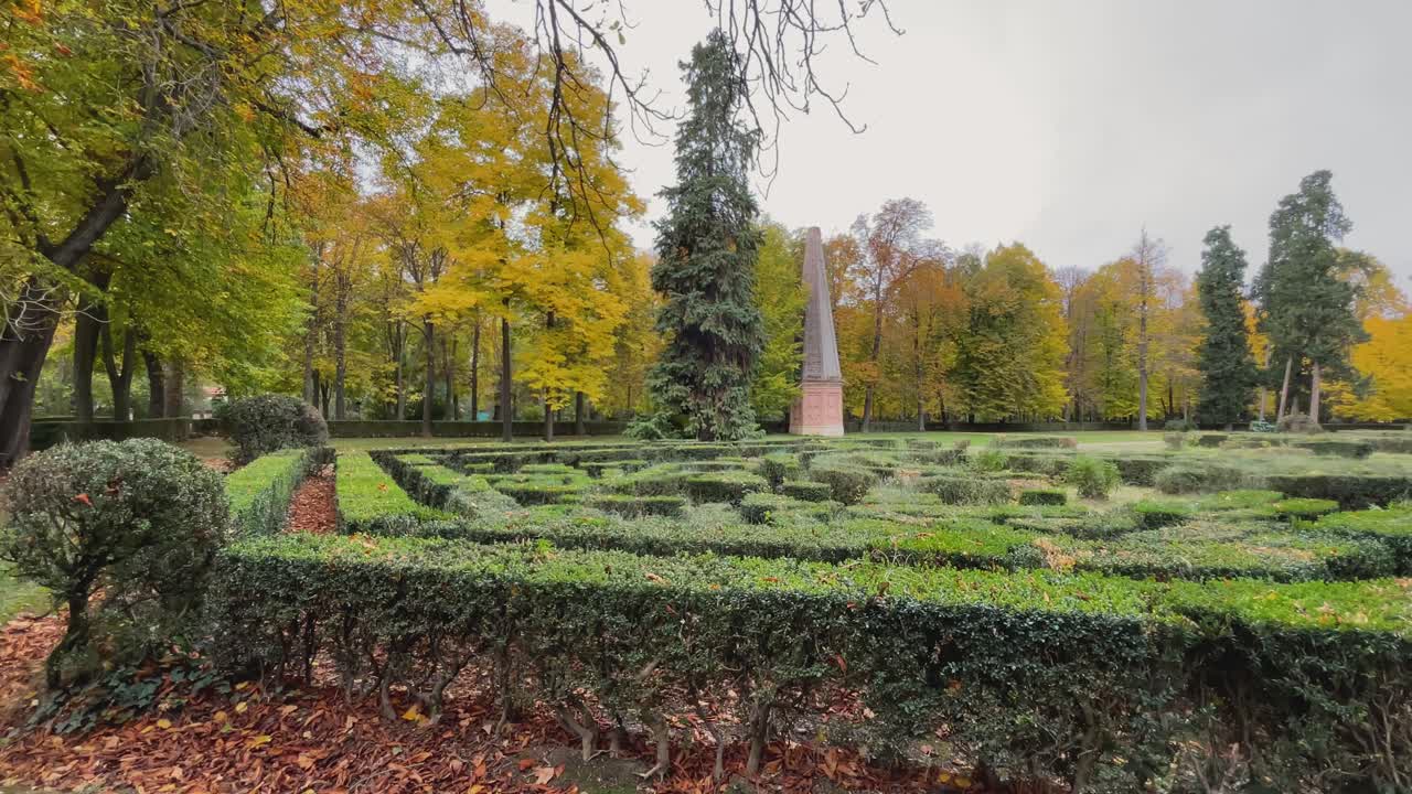 Balayage latéral au ralenti des Jardins Royaux d'Aranjuez. L'obélisque historique en brique se dresse en arrière-plan, derrière une barrière d'arbres, tandis que le jardin à la française, avec ses labyrinthes, occupe une position centrale.