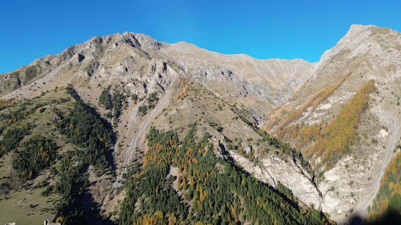 Drone captures rugged alpine mountains with rocky ridges and autumn-colored forests. Clear blue sky and November sunlight highlight the dramatic landscape near Serre Ponçon in the Hautes Alpes, France