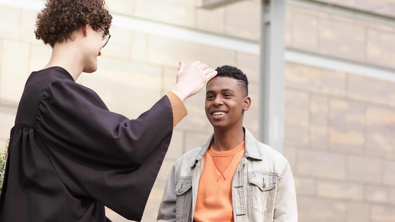 Diverse male friends snapping phone selfie at plaza, tucking mortarboard and diploma, shaking hands