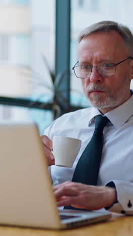 elderly man is sitting in a light room with big glass windows near computer with a cup of coffee. working on a latop in modern restaurant during coffee brake. vertical video