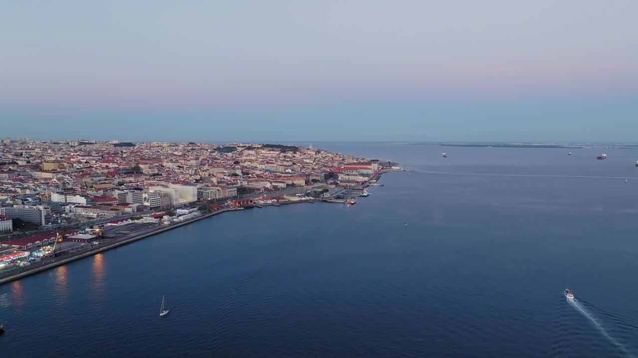 Splendide riprese aeree catturano lo skyline storico di Lisbona lungo l'estuario del fiume Tago al tramonto, con cieli pastello, acqua blu tranquilla, barche e luci della città.