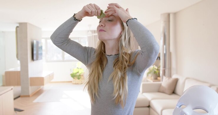 Donna che si porta le mani al viso, utilizzando un gua sha verde per massaggiare la mascella su un bancone in legno per la cura della pelle